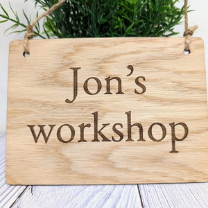 May include: A wooden sign with the text "Jon's workshop" engraved on it. The sign is hanging from a string and has two holes at the top.