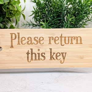 May include: A rectangular bamboo key tag with the engraved text "Please return this key." The tag has a silver key ring attached. The background features green foliage.