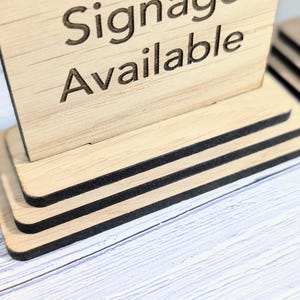 May include: A wooden sign with the text "Signage Available" on it. The sign is on a tiered stand with black trim.