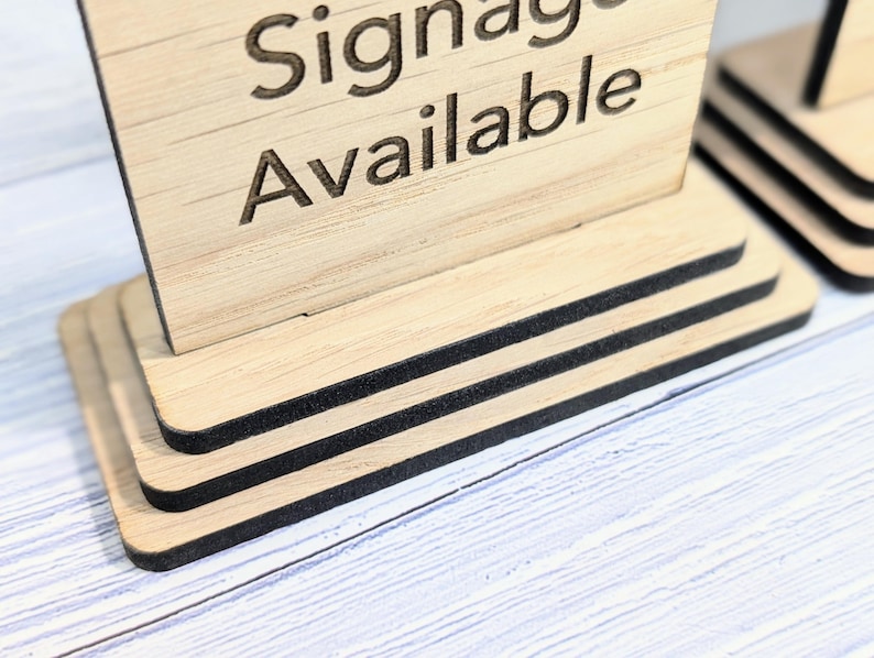 May include: A wooden sign with the text "Signage Available" on it. The sign is on a tiered stand with black trim.