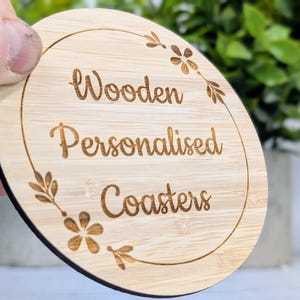 May include: A round, light-colored wooden coaster with the words "Wooden Personalised Coasters" engraved in a decorative font. The coaster has a floral design around the edge.