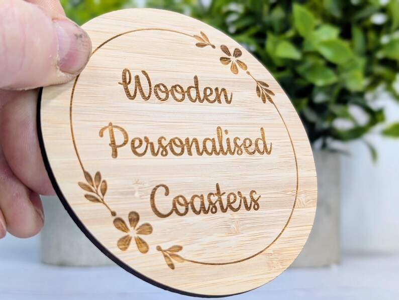 May include: A round, light-colored wooden coaster with the words "Wooden Personalised Coasters" engraved in a decorative font. The coaster has a floral design around the edge.