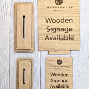 May include: Two wooden signs with the text "Cherry Grove Craft" and "Wooden Signage Available". The signs are made of light brown wood and have a natural grain pattern. The text is engraved into the wood and is black in color.