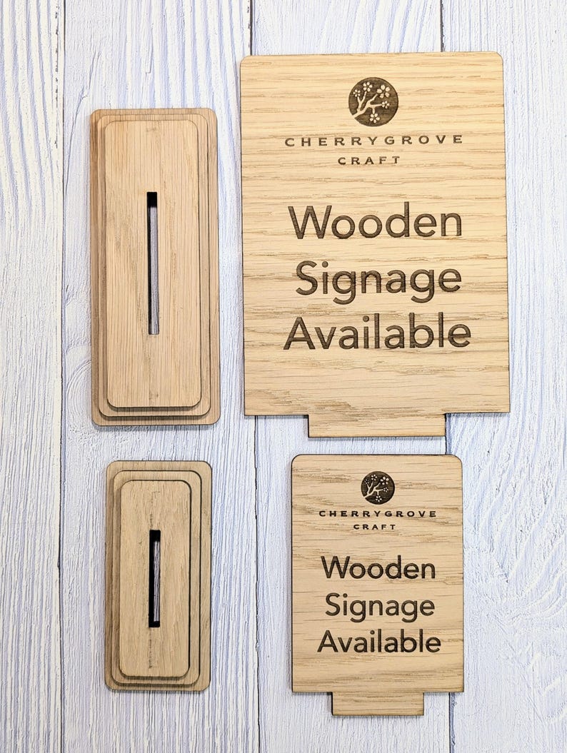 May include: Two wooden signs with the text "Cherry Grove Craft" and "Wooden Signage Available". The signs are made of light brown wood and have a natural grain pattern. The text is engraved into the wood and is black in color.