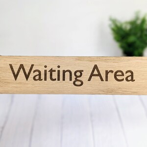 May include: A wooden sign with the text "Waiting Area" engraved on it. The sign is light brown and has two small holes on either side for hanging.