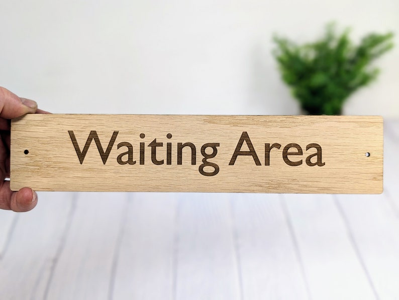 May include: A wooden sign with the text "Waiting Area" engraved on it. The sign is light brown and has two small holes on either side for hanging.