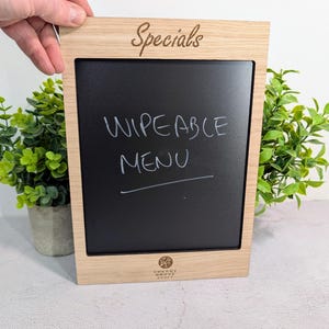 May include: A wooden framed chalkboard with the word "Specials" engraved at the top. The black wipeable surface displays the words "WIPEABLE MENU" in white. The frame is held by a hand, with greenery in the background.