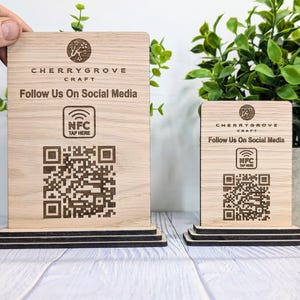 以下が含まれることがあります： 「CHERRYGROVE CRAFT」と「Follow Us On Social Media」と書かれた木製の看板が2つ。それぞれの看板にはQRコードと「NFC TAP HERE」の文字が書かれています。