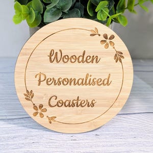 May include: A round, light-colored wooden coaster with the words "Wooden Personalised Coasters" engraved in a circular design. Floral and leaf accents surround the text. The coaster is made of bamboo.