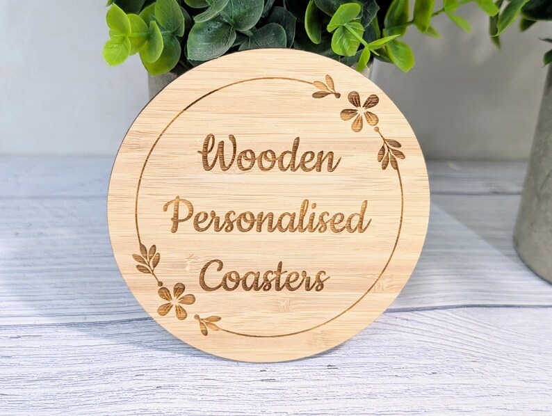 May include: A round, light-colored wooden coaster with the words "Wooden Personalised Coasters" engraved in a circular design. Floral and leaf accents surround the text. The coaster is made of bamboo.