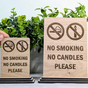 May include: Two wooden signs with black text and symbols. The larger sign reads "No Smoking No Candles Please". The smaller sign reads "No Smoking No Candles Please". Both signs have a no smoking symbol and a no candle symbol.