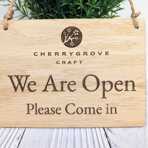 May include: A wooden sign with a cherry blossom logo and the text "Cherrygrove Craft" above the text "We Are Open" and "Please Come In".