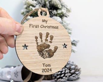Op de afbeelding: Een houten kerstversiering met een handafdruk, sterren en de tekst "My First Christmas Tom 2024".