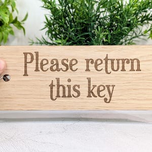 May include: A rectangular wooden key tag with the engraved text "Please return this key." The tag is light brown with a silver key ring attached. The background includes green plants and a white surface.