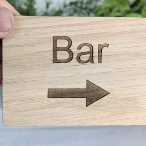 Könnte beinhalten: Ein Holzschild mit dem Wort "Bar" und einem Pfeil, der nach rechts zeigt. Das Schild ist hellbraun mit einer Holzmaserung.