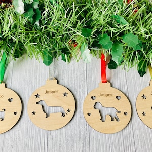 May include: Four wooden Christmas ornaments with dog silhouettes and names. The ornaments are in the shape of a ball and have a ribbon attached. The names on the ornaments are Coco, Jasper, Jasper, and Jasper. The dog silhouettes are a Yorkshire Terrier, a Dachshund, a Labradoodle, and a paw print.