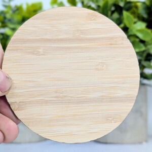 May include: A round, light-colored bamboo coaster with a natural wood grain pattern. The coaster is held by a hand, with green foliage and a concrete pot in the background. The coaster is approximately 4 inches in diameter.
