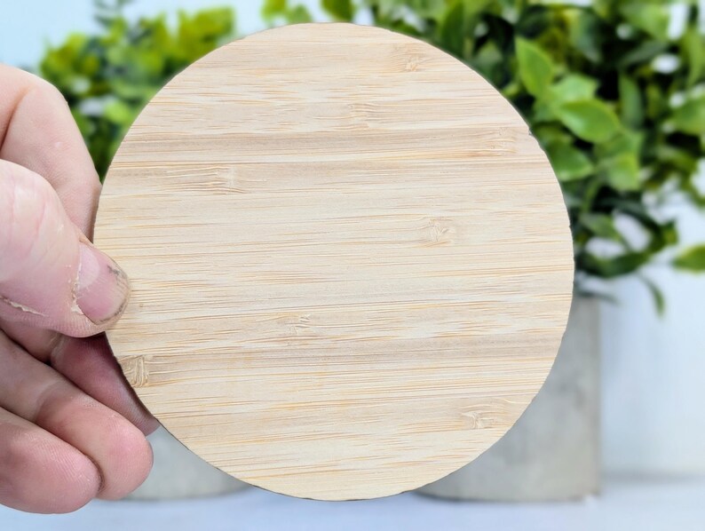 May include: A round, light-colored bamboo coaster with a natural wood grain pattern. The coaster is held by a hand, with green foliage and a concrete pot in the background. The coaster is approximately 4 inches in diameter.