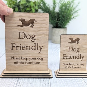 May include: Two wooden signs with the text "Dog Friendly" and a silhouette of a dog. The signs are on a wooden stand and have the text "Please keep your dog off the furniture".