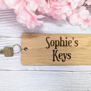 May include: A wooden key holder with the text "Sophie's Keys" engraved on it. The key holder is light brown and has a key ring attached to it. The key holder is sitting on a white wooden surface.