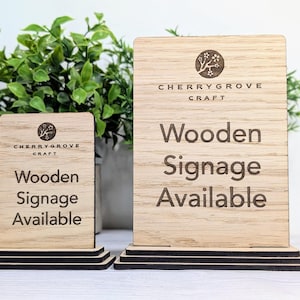 May include: Two wooden signs with a black stand. The signs are made of light brown wood and have the text "Cherrygrove Craft" and "Wooden Signage Available" engraved on them.