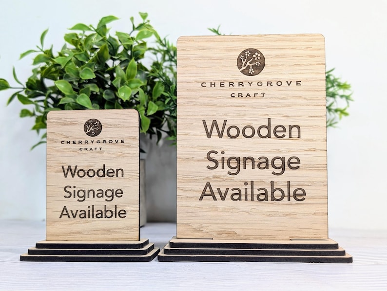 May include: Two wooden signs with a black stand. The signs are made of light brown wood and have the text "Cherrygrove Craft" and "Wooden Signage Available" engraved on them.