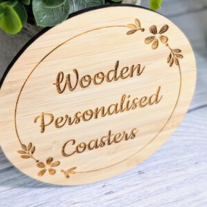 May include: A round, light-colored wooden coaster with the words "Wooden Personalised Coasters" engraved in a decorative font. The coaster has a black edge and is adorned with floral accents.