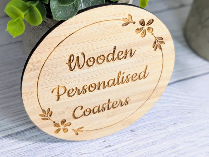 May include: A round, light-colored wooden coaster with the words "Wooden Personalised Coasters" engraved in a decorative font. The coaster has a black edge and is adorned with floral accents.