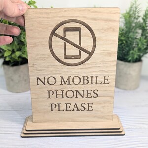 May include: Wooden sign with a red circle and diagonal line through a phone icon. The sign reads "No Mobile Phones Please".