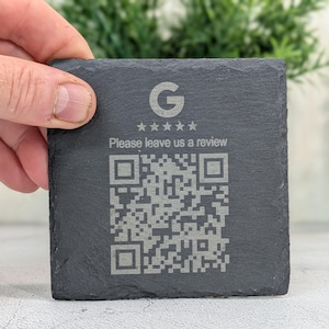 May include: A square, dark grey slate coaster featuring the Google logo, four stars, the text "Please leave us a review," and a QR code. The coaster is held by a hand.