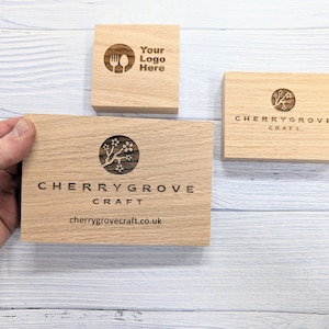 Puede incluir: Tres letreros de madera con texto grabado. El letrero superior dice "Your Logo Here" con un símbolo de tenedor y cuchillo. El letrero del medio dice "CHERRY GROVE CRAFT" con un árbol de cerezo en flor estilizado. El letrero inferior dice "CHERRY GROVE CRAFT" con el sitio web "cherrygrovecraft.co.uk".