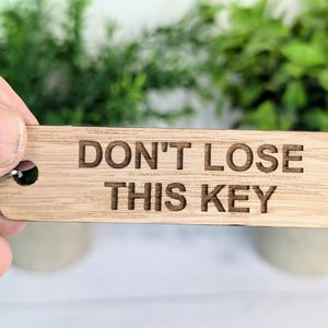 Könnte beinhalten: Ein Holzschlüsselanhänger mit dem eingravierten Text "DON'T LOSE THIS KEY". Der Schlüsselanhänger ist an einem silbernen Schlüsselring befestigt.