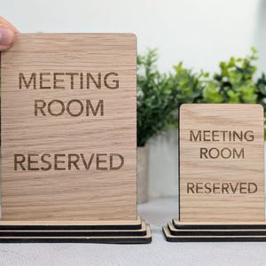 May include: Two wooden signs, one held by a hand, with the text "MEETING ROOM RESERVED". The signs are light brown with a wood grain pattern and sit on a black base. Green plants are in the background.