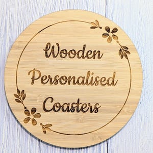 May include: A round, wooden coaster with the words "Wooden Personalised Coasters" engraved in a decorative font. The coaster has a floral design around the edge.