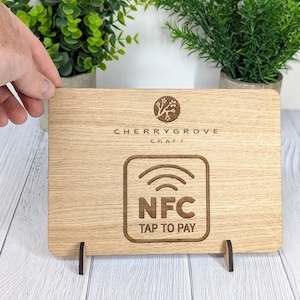 May include: Wooden sign with a brown woodgrain finish. The sign has a white logo with a tree and the text "Cherrygrove Craft". Below the logo is a white square with a stylized image of a wireless signal and the text "NFC Tap to Pay". The sign is on a white wooden stand.