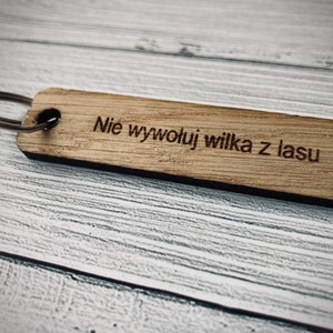 May include: A wooden keychain with a silver ring and the text "Nie wywołuj wilka z lasu" engraved on it.