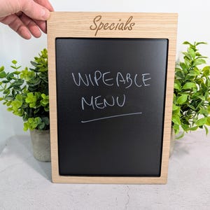 May include: A rectangular chalkboard with a light wood frame. The word "Specials" is scripted at the top. The black surface displays the words "WIPEABLE MENU" in white. The chalkboard is held by a hand, with green plants in the background.
