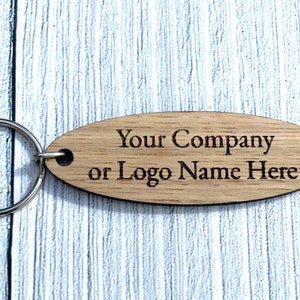 Könnte beinhalten: Ovaler Holzschlüsselanhänger mit einem silbernen Schlüsselring. Der Schlüsselanhänger ist mit dem Text "Your Company or Logo Name Here" graviert.