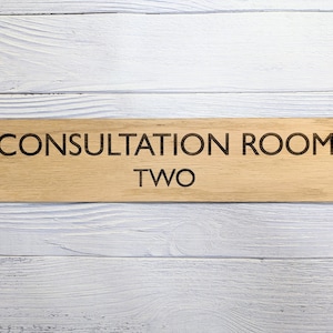 May include: Wooden sign with the text "Consultation Room Two" in black lettering on a light brown background.