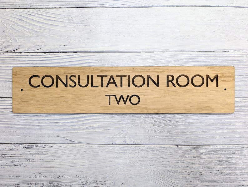 May include: Wooden sign with the text "Consultation Room Two" in black lettering on a light brown background.