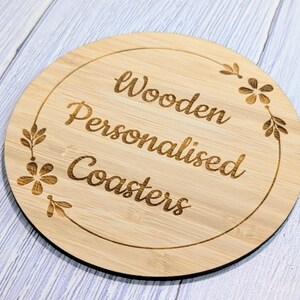 May include: A round, light-brown wooden coaster with the words "Wooden Personalised Coasters" engraved in a cursive font. The coaster has a decorative floral border. The coaster is on a light blue and white surface.