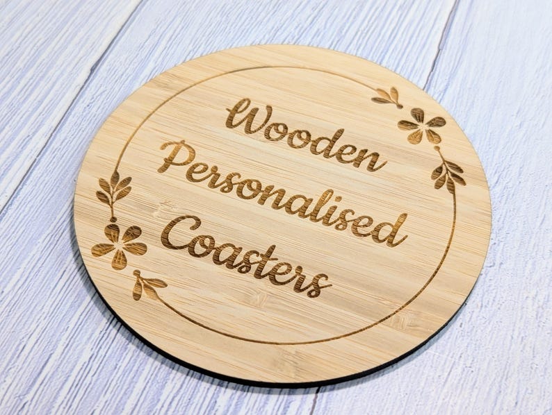May include: A round, light-brown wooden coaster with the words "Wooden Personalised Coasters" engraved in a cursive font. The coaster has a decorative floral border. The coaster is on a light blue and white surface.