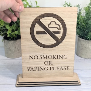 May include: Wooden sign with a no smoking symbol and the text "NO SMOKING OR VAPING PLEASE" in a dark brown colour. The sign is made of light-coloured wood and has a rectangular shape with a base. The sign is held by a hand.