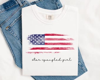 May include: White t-shirt with a distressed American flag design in red, white, and blue. The text "star spangled girl" is printed below the flag. The shirt is laid on top of blue denim jeans.
