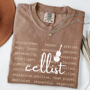 May include: A brown Comfort Colors t-shirt with the word "cellist" in white script, surrounded by descriptive words. A silver necklace with a pendant is draped across the shirt. The shirt is size small.