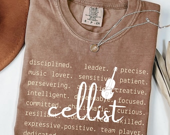Peut inclure: Un t-shirt Comfort Colors marron avec le mot "cellist" en écriture blanche, entouré de mots descriptifs. Un collier argenté avec un pendentif est drapé sur le t-shirt. Le t-shirt est de taille small.
