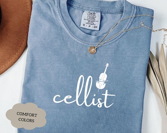 Peut inclure: Un t-shirt bleu clair avec le mot "cellist" écrit en lettres cursives blanches. Un contour blanc d'un violoncelle est au-dessus du mot. Le t-shirt est fabriqué par Comfort Colors.