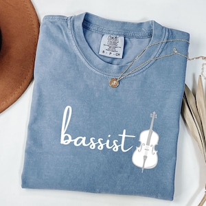 Puede incluir: Una camiseta azul con la palabra "bajista" en escritura blanca y un gráfico de violonchelo blanco. Un collar dorado con un colgante está drapeado sobre la camiseta. La camiseta es de Comfort Colors.