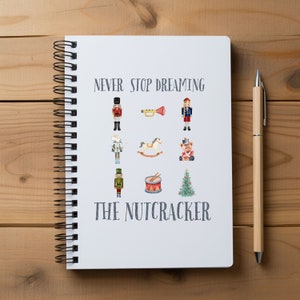 Puede incluir: Una libreta de espiral blanca con el texto "NEVER STOP DREAMING THE NUTCRACKER" impreso en la portada. La portada también presenta ilustraciones de un cascanueces, un caballito de balancín, un tambor, un árbol de Navidad y una trompeta.