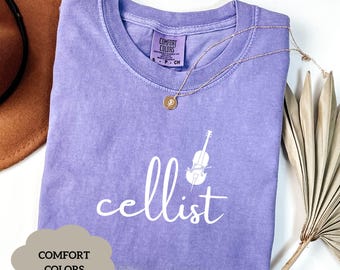 Peut inclure: Un t-shirt violet avec le mot "cellist" et un contour blanc d'un violoncelle. Le t-shirt est fabriqué par Comfort Colors.